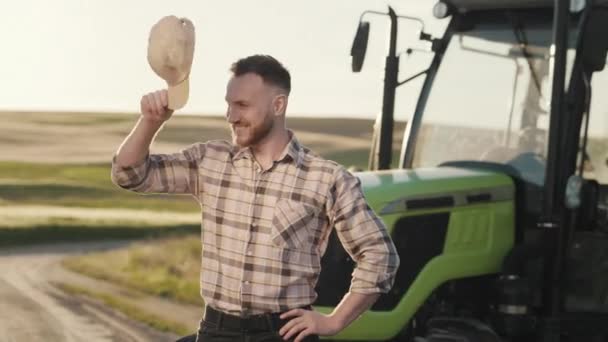 Ein junger bärtiger Bauer setzt sich eine Mütze auf und hält seine Hände am Gürtel. Er lächelt und blickt in die Kamera. Er steht mitten auf einem Feld neben einem Traktor. Die Sonne scheint. 4K — Stockvideo