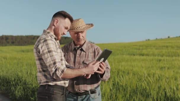 Um homem está a mostrar informações a um velho agricultor numa tábua. Ele está apontando para o campo. Estão a discutir a colheita. 4K — Vídeo de Stock