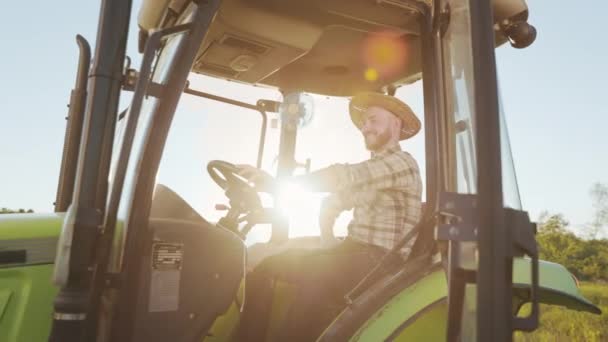Ein junger Landwirt sitzt am Steuer eines Traktors. Er blickt in die Kamera und lächelt. Er trägt einen Strohhut. Sonnenuntergang im Hintergrund. 4K — Stockvideo