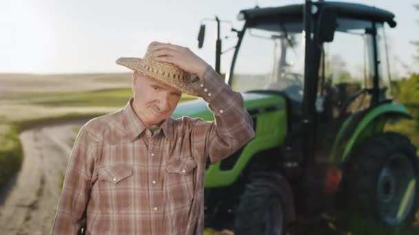 Stary farmer zakłada słomkowy kapelusz i patrzy w kamerę. Stoi na środku pola w pobliżu traktora. Zachód słońca w tle. 4K — Wideo stockowe