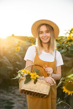 Bir kadın elinde bir sepet çiçekle ayçiçeği tarlasında dikiliyor. Gülümsüyor ve kameraya bakıyor. Gün batımı.