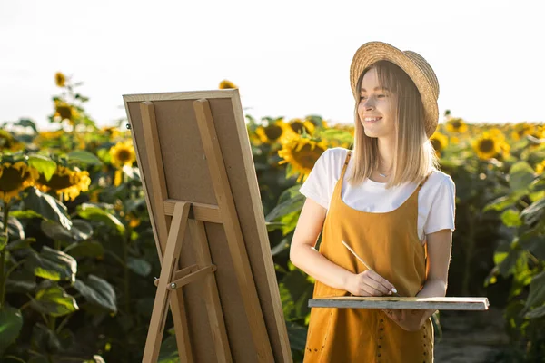 Bir kadın ayçiçeği tarlasında duruyor ve resim çiziyor. Hasır şapka takıyor. Tuval bir sehpanın üzerinde duruyor. O bir fırça ve palet tutuyor. Gülümsüyor..