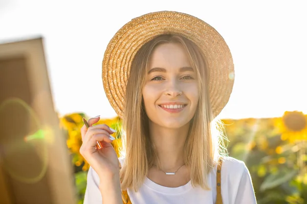 Ayçiçeği tarlasında dikilip kameraya bakan bir kadının portresi. Tuval bir sehpanın üzerinde duruyor. Gülümsüyor ve elinde fırça tutuyor..