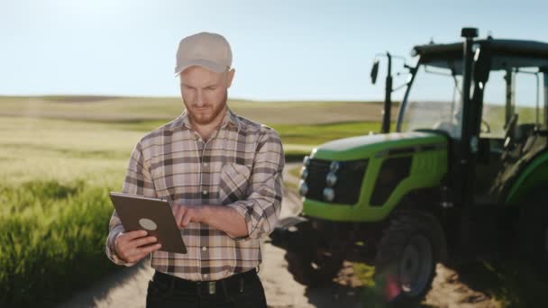 Um jovem agricultor barbudo está num campo perto de um tractor. Ele está a trabalhar num tablet. Ele está a usar um boné. Pôr-do-sol ao fundo. 4K — Vídeo de Stock