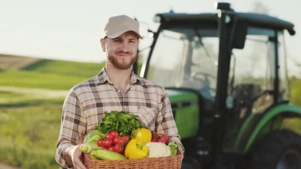 Фермер стоит возле поля и держит корзину с овощами с поля. Он улыбается. Он поднимает голову и смотрит в камеру. Трактор на заднем плане. 4K — стоковое видео