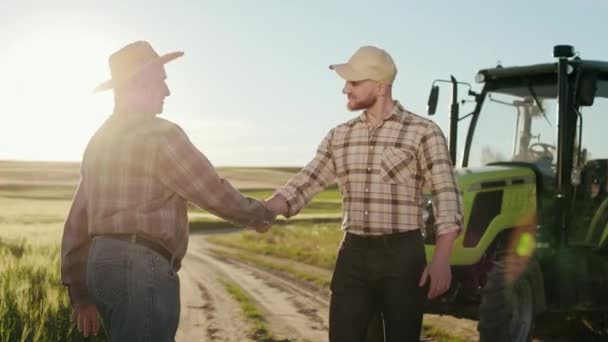 Два фермера встречаются и пожимают друг другу руки. Они улыбаются и разговаривают. За ними есть трактор. Солнце светит ярко. 4K — стоковое видео