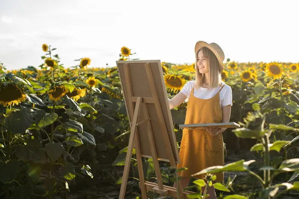 Bir kadın ayçiçeği tarlasında duruyor ve resim çiziyor. Hasır şapka takıyor. Tuval bir sehpanın üzerinde duruyor. Gülümsüyor..