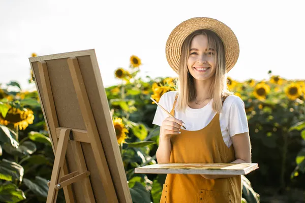 Bir kadın ayçiçeği tarlasında duruyor ve resim çiziyor. Tuval bir sehpanın üzerinde duruyor. Gülümsüyor ve kameraya bakıyor. Elinde bir fırça ve palet tutuyor..