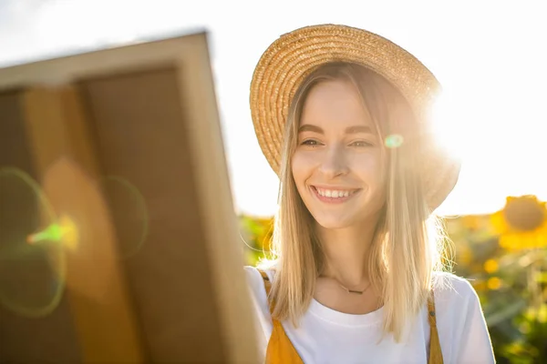 Ayçiçeği tarlasında duran ve resme bakan bir kadının portresi. Tuval bir sehpanın üzerinde duruyor. Gülümsüyor ve elinde fırça tutuyor..
