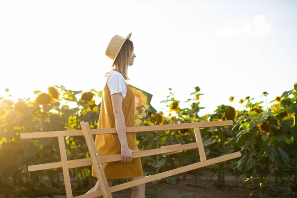 Genç bir kadın sehpa ve tablo taşıyor. Ayçiçeği tarlasında yürüyor. Doğada sanatçı.