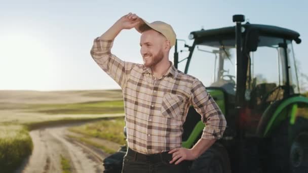 Um jovem agricultor barbudo está a colocar um boné e a manter as mãos no cinto. Ele está sorrindo. Ele está no meio de um campo perto de um trator. Pôr-do-sol ao fundo. 4K — Vídeo de Stock