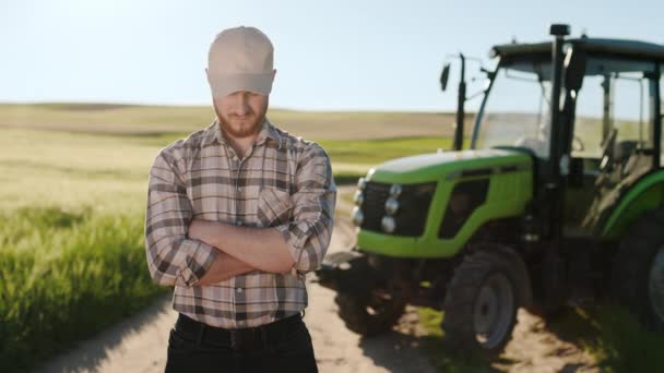 Um jovem agricultor barbudo está num campo perto de um tractor. Ele está levantando a cabeça e olhando para a câmera. Ele está a usar um boné. Pôr-do-sol ao fundo. 4K — Vídeo de Stock