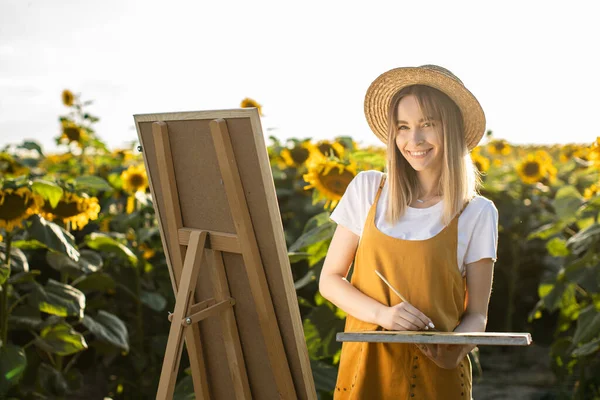 Bir kadın ayçiçeği tarlasında duruyor ve resim çiziyor. Hasır şapka takıyor. Tuval bir sehpanın üzerinde duruyor. Gülümsüyor ve kameraya bakıyor..