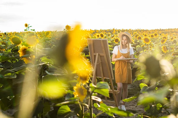 Bir kadın ayçiçeği tarlasında duruyor ve resim çiziyor. Tuval bir sehpanın üzerinde duruyor. O gülümsüyor ve resme bakıyor. Ayçiçekleri ön planda.