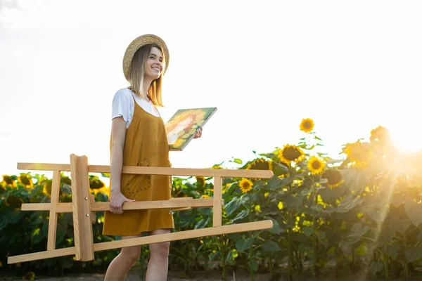 Genç bir kadın sehpa ve tablo taşıyor. Ayçiçeği tarlasında yürüyor. Gülümsüyor..