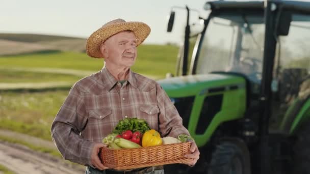 Egy öreg farmer áll egy traktornál a mezőn, és a kamerába néz. Egy kosár zöldséget és füvet tart a kezében. Kalap van rajta. A kamera forog körülötte. 4K — Stock videók