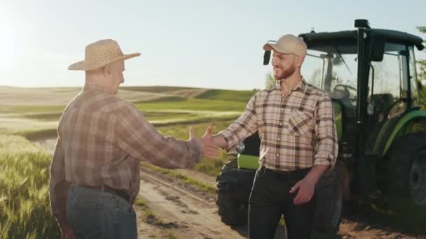 Dois agricultores estão se reunindo e apertando as mãos. Estão a sorrir. Há um tractor atrás deles. O sol está brilhando intensamente. 4K — Vídeo de Stock