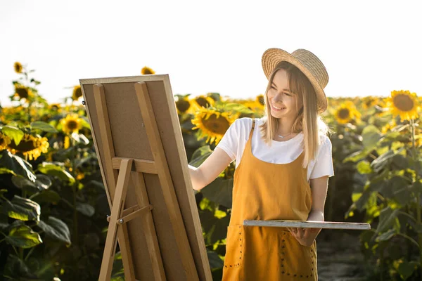 Bir kadın ayçiçeği tarlasında duruyor ve resim çiziyor. Hasır şapka takıyor. Tuval bir sehpanın üzerinde duruyor. O gülümsüyor. Güneş ışıldıyor..