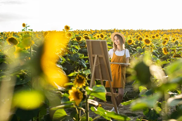 Bir kadın ayçiçeği tarlasında duruyor ve resim çiziyor. Tuval bir sehpanın üzerinde duruyor. O gülümsüyor. Ayçiçekleri ön planda.