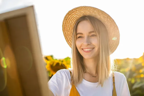 Ayçiçeği tarlasında duran ve resme bakan bir kadının portresi. Tuval bir sehpanın üzerinde duruyor. Gülümsüyor..