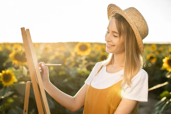 Ayçiçeği tarlasında duran ve resme bakan bir kadının portresi. Tuval bir sehpanın üzerinde duruyor. Elinde bir fırça tutuyor ve tuvale resim çiziyor..
