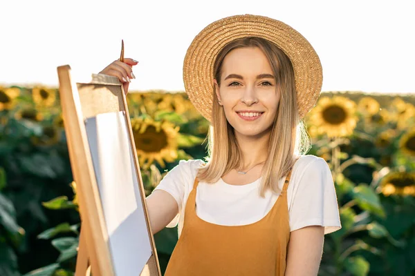 Gülümseyen bir kadının portresi. O, ayçiçeklerinin arasında bir sehpanın yanında duruyor. O, kameraya bakıyor. Elinde fırça tutuyor..
