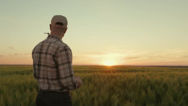 Ein junger Bauer steht mitten auf einem Feld und steuert eine Drohne, die ihn überfliegt. Er vermessen das Feld mit einer Drohne. Sonnenuntergang im Hintergrund. 4K — Stockvideo