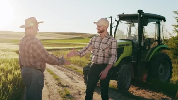 O homem está se aproximando do velho fazendeiro e eles estão apertando as mãos. Estão a sorrir. Há um tractor atrás deles. O sol está brilhando intensamente. 4K — Vídeo de Stock