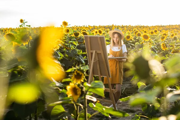 Bir kadın ayçiçeği tarlasında duruyor ve resim çiziyor. Tuval bir sehpanın üzerinde duruyor. O bir fırça ve palet tutuyor. Ayçiçekleri ön planda.