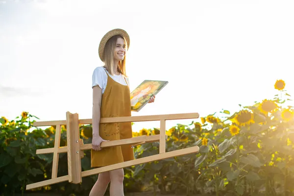 Genç bir kadın sehpa ve tablo taşıyor. Ayçiçeği tarlasında yürüyor..