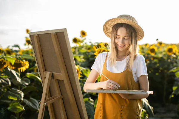 Bir kadın ayçiçeği tarlasında duruyor ve resim çiziyor. Hasır şapka takıyor. Tuval bir sehpanın üzerinde duruyor. Elinde bir fırça ve palet tutuyor..