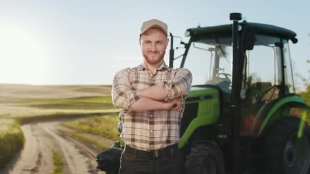 Um jovem agricultor está cruzando os braços e olhando para a câmera. Ele está sorrindo. Ele está no meio de um campo perto de um trator. Pôr-do-sol ao fundo. 4K — Vídeo de Stock