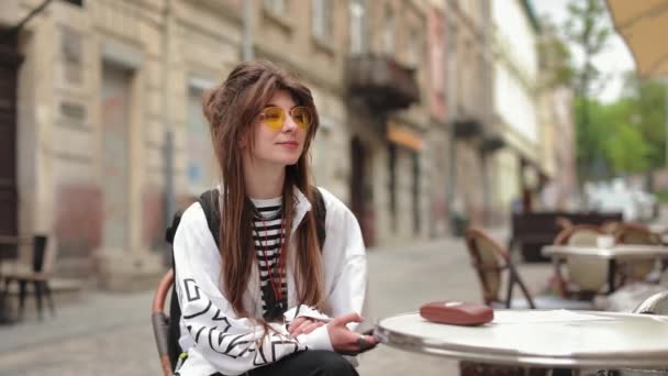 Muchacha hipster retrato que está sentado en un café en la vieja calle. Ella está mirando a su alrededor y sonriendo. Lleva gafas amarillas. 4K — Vídeo de stock