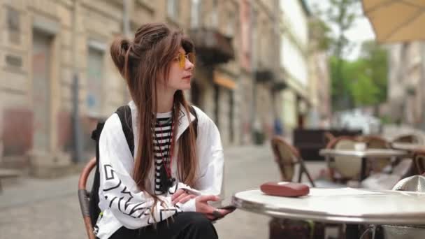 Muchacha hipster retrato que está sentado en un café en la vieja calle. Ella está mirando a su alrededor y sonriendo. Ella sostiene un teléfono inteligente en sus manos. Lleva gafas amarillas. 4K — Vídeos de Stock