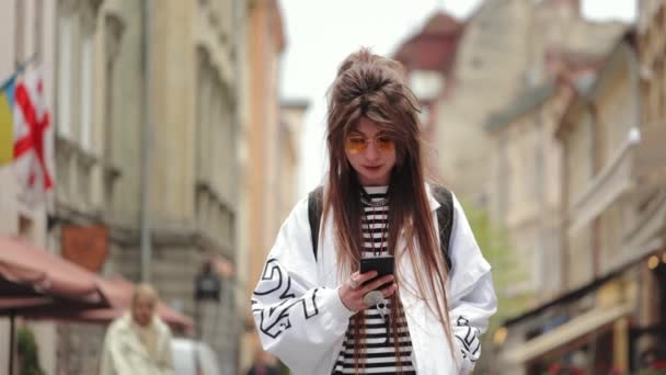 Chica Hipster está de pie en el centro de la ciudad. Ella está enviando mensajes en su teléfono inteligente. Está levantando la cabeza y mirando a la cámara. Lleva gafas amarillas. 4K — Vídeos de Stock