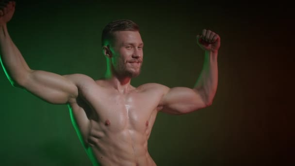 Ένας αθλητής με γυμνό σώμα στέκεται σε μια στάση για να δείξει τους δικέφαλους. Χαμογελάει και κοιτάζει την κάμερα. Ένα πορτοκαλί και πράσινο φως λάμπει πάνω του. 4K — Αρχείο Βίντεο