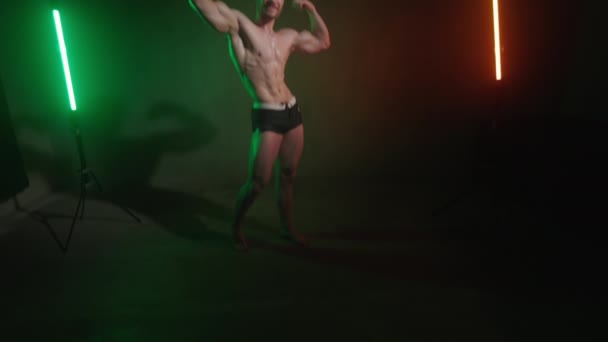Un deportista con un cuerpo desnudo posando y mostrando bíceps. La cámara está subiendo. Una luz naranja y verde brilla sobre él. 4K — Vídeo de stock