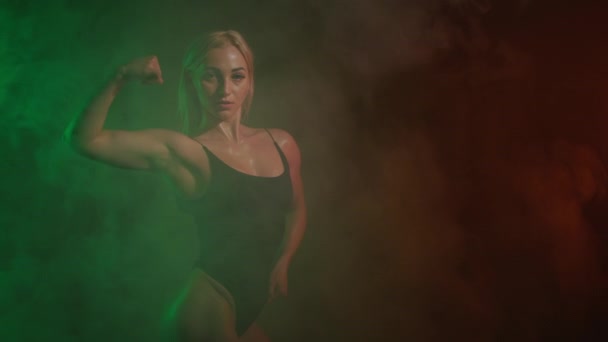 Fitness modelka v teplákovém obleku pózuje. Dívá se do kamery a ukazuje biceps. Všude kolem ní je kouř. Oranžové a zelené světlo na ni svítí. 4K — Stock video
