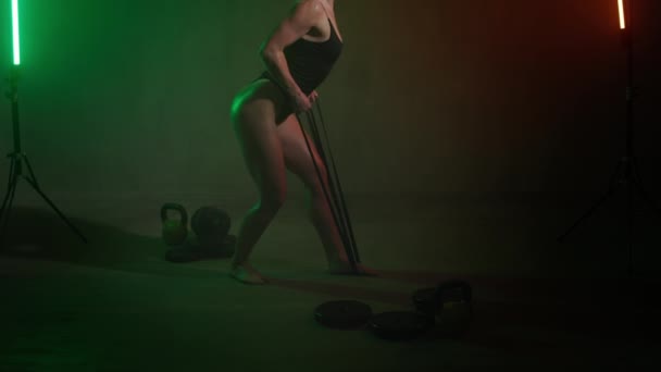 Een fitnessmodel in een bodysuit poseert. Ze doet rugoefeningen met verzetsbanden. Er zijn halters en kettlebells bij haar in de buurt. Een oranje en groen licht schijnt op haar. 4K — Stockvideo