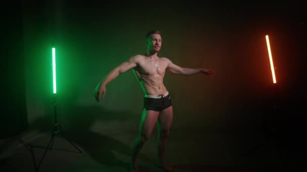 Спортсмен с голым телом поднимает руки и стоит в позе, чтобы показать свои мускулы. Он улыбается. Камера уменьшает масштаб. Ему светит оранжевый и зеленый свет. 4K — стоковое видео