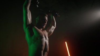 Bir sporcu başının üstüne dambıl bastırıyor. O, omuzunda ağır bir set yapıyor. Turuncu ve yeşil ışık onun üzerinde parlıyor. 4K