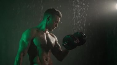 Sporcu duşta duruyor ve pazı egzersizi yapıyor. O, dambılları teker teker kaldırıyor. Vücudundan su akıyor. Üzerinde yeşil ışık parlıyor. 4K
