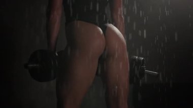 Fitness modeli duşta duruyor ve dambılları tutuyor. O, kalçalarını gösteriyor. Seksi bir vücut. Vücudundan su akıyor. 4K