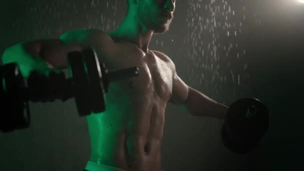 El atleta está de pie en la ducha y realizando un ejercicio de hombros. Está agitando pesas a un lado. El agua corre por su cuerpo. Una luz verde está brillando sobre él. 4K — Vídeo de stock