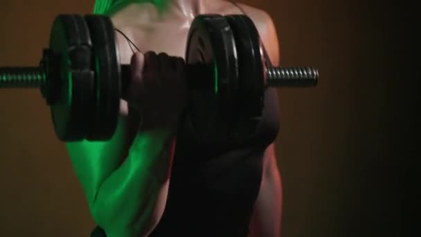 Träningsmodellen står och tränar biceps. Hon böjer armen med en hantel. Kameran fokuserar på hanteln. Ett orange och grönt ljus lyser på henne. 4K 50fps — Stockvideo