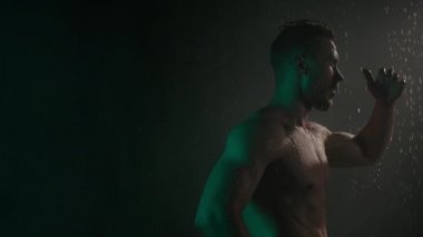 Sportif bir adam saçlarını eliyle düzeltiyor. O, vücudunu gösteriyor. Vücudundan su akıyor. Yeşil ışık onun üzerinde parlıyor. 4K
