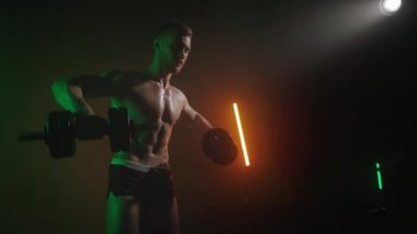 Sporcu halterleri yana doğru sallar. O, omuzunda ağır bir set yapıyor. Önüne bakıyor. Turuncu ve yeşil ışık onun üzerinde parlıyor. 4K