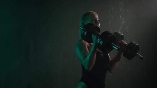 O modelo de fitness está de pé no chuveiro e fazendo um exercício de bíceps. Ela está dobrando os braços com halteres. A água está a escorrer pelo corpo. Uma luz verde está brilhando sobre ela. 4K — Vídeo de Stock