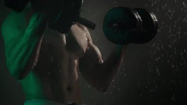 Sporcu duşta duruyor ve pazı egzersizi yapıyor. O, halterleri kaldırıyor. Kamera vücuduna odaklanıyor. Vücudundan su akıyor. Üzerinde yeşil ışık parlıyor. 4K