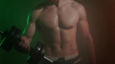 Sporcu ayakta duruyor ve pazı egzersizi yapıyor. O, dambılları teker teker kaldırıyor. Kamera dambıllara odaklanıyor. Onunkinde turuncu ve yeşil ışık parlıyor. 4K 50fps